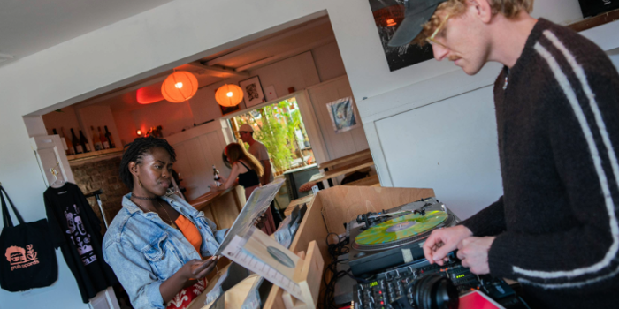A person stands at a wooden record counter browsing vinyl records, while another operates DJ equipment with turntables and a mixer. Headphones and a colourful record sleeve are visible on the counter. The setting appears to be a cosy shop or café with white walls, hanging orange lights, shelves with bottles, and a tote bag displayed on the wall. A doorway in the background reveals more people and greenery outside.