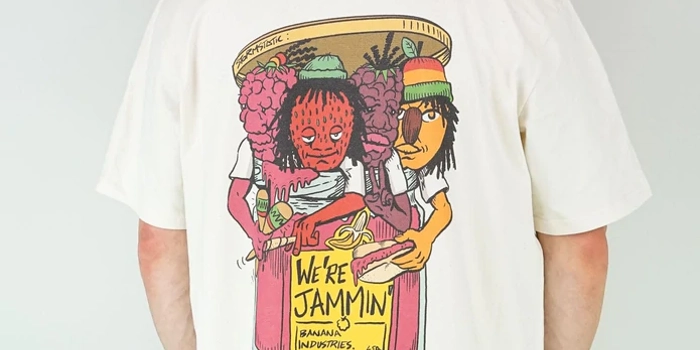 A man stands with his back to the camera, wearing a baseball bat, a white t-shirt with a colourful back print and green trousers. The design on the shirt is four anthropomorphic fruits busting out of a jam jar. They look like members of a band. The yellow label on the jar reads 'We're jammin''.