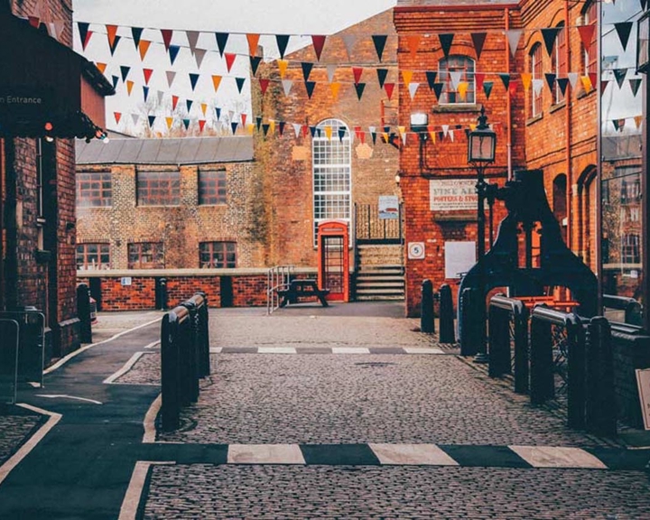 Cobblestone courtyard surrounded by historic brick buildings, decorated with strings of triangular bunting in red, white, and blue. A traditional red phone box stands near a large arched window at the far end. Signs on the right indicate museum entrance, admissions, shop, café, and toilets.