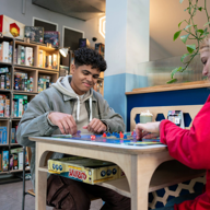 Two people are sat at a table, in a cafe, playing a board game and drinking coffees. Behind them is shelving stacked full of all different board games.