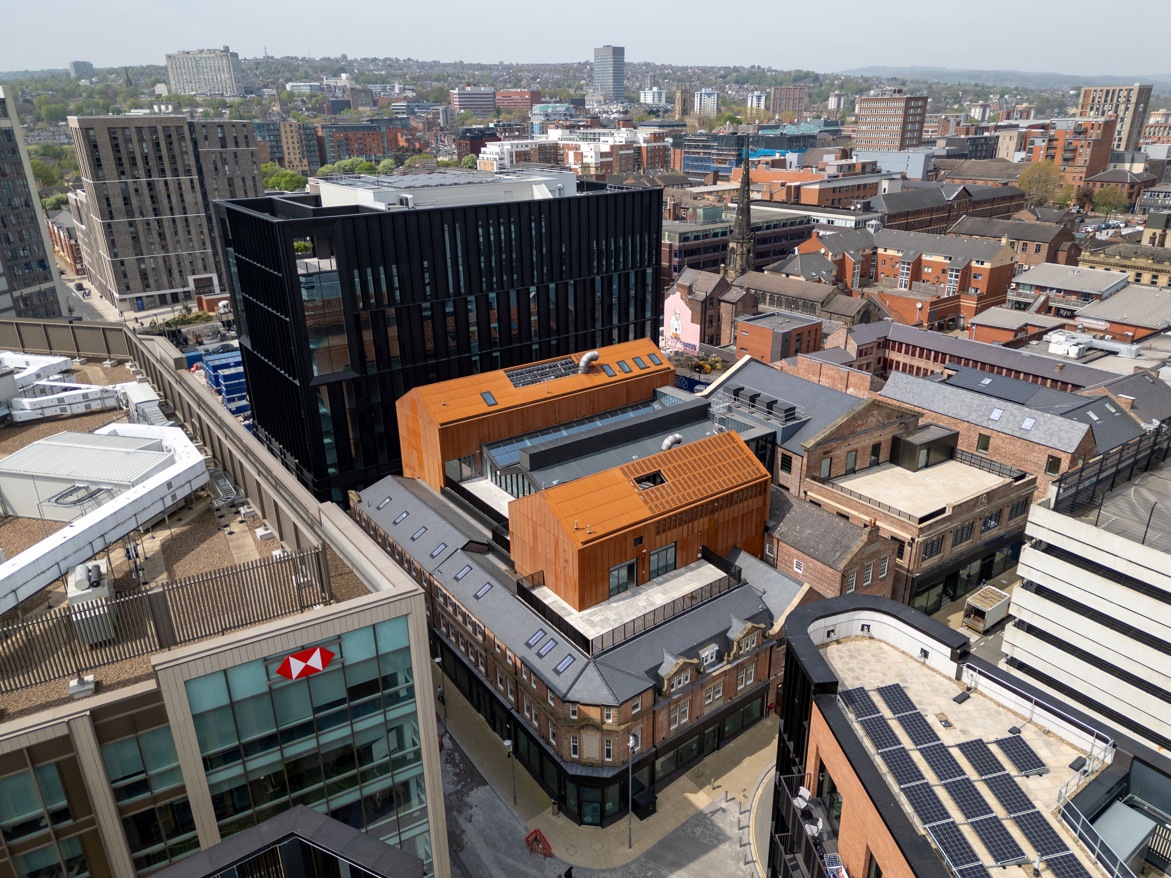 Ariel shot of Cambridge Street Collective. Aerial view of an urban area featuring modern and historic architecture. Prominent buildings include a black glass high-rise and orange-clad structures surrounded by older brick buildings. The scene shows a mix of commercial and residential properties, with solar panels visible on one rooftop and an HSBC building in the foreground. The background includes a sprawling city-scape with trees and hills under a clear sky