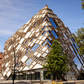 A modern, pyramid-shaped building with a striking geometric design featuring a mix of glass panels and wooden lattice structures. The facade reflects the blue sky, and the building is surrounded by trees and a few older brick buildings on the left. The entrance area has large glass doors, and the street in front includes signage and a lamppost.