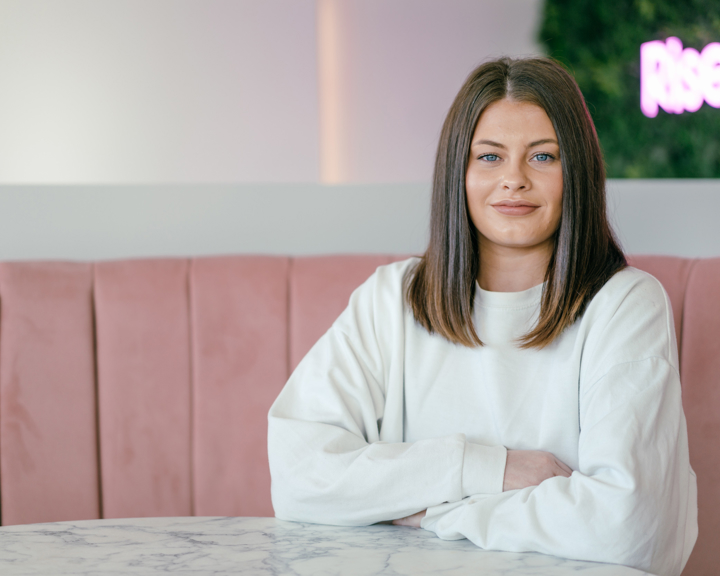 A person wearing a plain white long-sleeve top is seated at a round marble table with arms resting on the surface. The background features a pink cushioned booth and a wall with greenery and partially visible neon text. The setting appears modern and stylish, likely in a café or casual dining space.