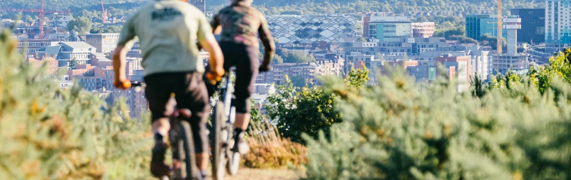 Two people are mountain biking along a dirt track that is flanked with heather on both sides. In the distance the Sheffield skyline is visible.