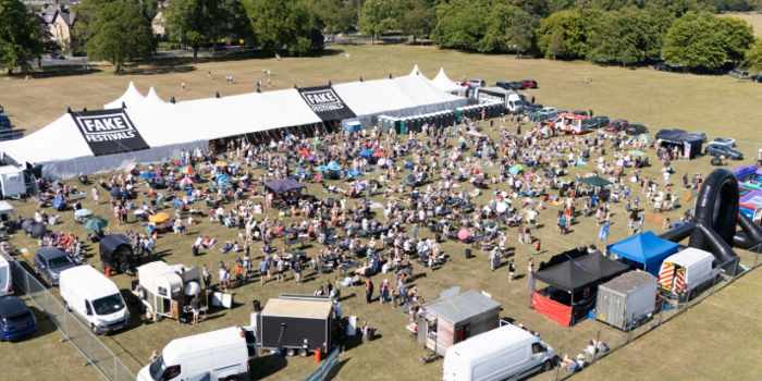 A large outdoor music festival set in an open grassy field with a white marquee tent labeled “FAKE FESTIVALS.” Crowds of people are gathered in front of the tent, sitting on blankets or standing near the stage area. Surrounding the festival are food stalls, trucks, and inflatable attractions, with trees and countryside visible in the background under a clear blue sky.