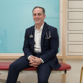 A person wearing a dark navy suit with a white dress shirt, seated on a red cushioned examination table in a clinical setting. A stethoscope hangs around the neck, suggesting a medical professional. The background includes a light blue frosted glass panel and a section of white drawers on the right side.