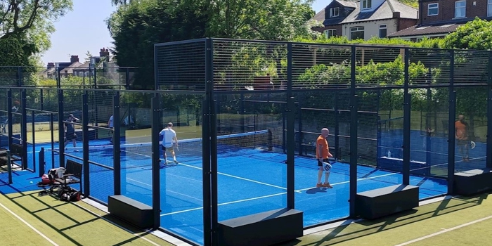 Outdoor padel courts with bright blue playing surfaces enclosed by glass and metal fencing, set within a larger green tennis court area. Several players are on the padel courts, holding rackets and playing under clear blue skies. Surrounding the courts are tall trees and residential houses, creating a leafy suburban backdrop. Sports bags and equipment are placed near the court entrance.