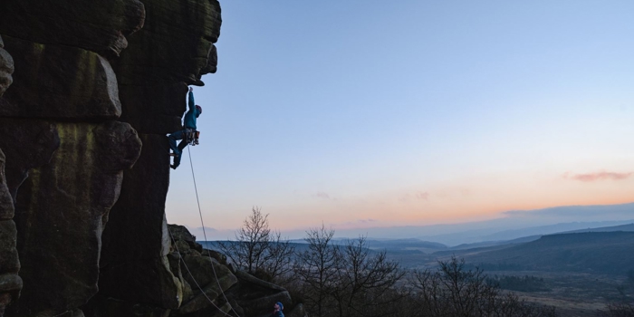 A person is rock climbing on a steep cliff face at dusk, secured by a rope held by another person standing below. The sky is clear with a gradient from blue to orange near the horizon, and the landscape in the background features rolling hills and sparse trees.
