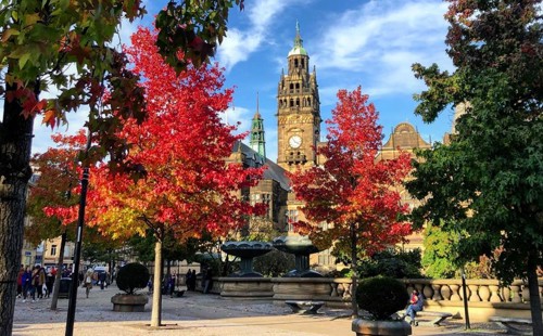 City square with vibrant autumn trees in shades of red and orange, framing a historic building with a tall clock tower in the background. The scene includes ornate fountains, stone balustrades, and people walking around under a bright blue sky with scattered clouds.