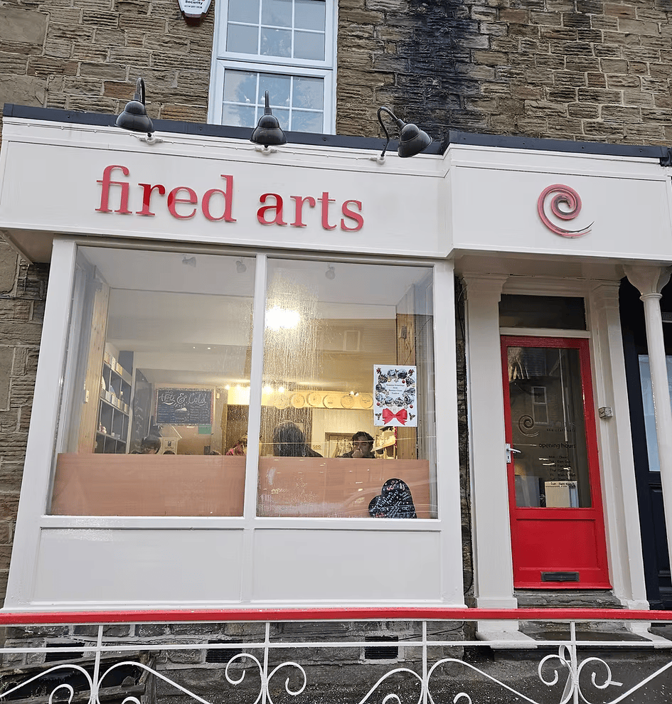 Exterior of Fired Arts ceramics studio with white frontage, red door, and large front window showing the studio interior.