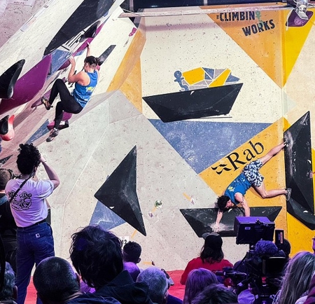 Two climbers wearing blue tops compete on an indoor bouldering wall with large black and purple holds. The wall features bold geometric panels in yellow, white, and blue with visible event branding, including “Rab” and “Climbin Works.” A crowd of spectators and camera operators watch from the foreground under bright stage lighting.