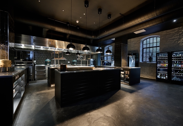 Modern professional kitchen with dark flooring and black cabinetry. The space features large stainless steel counters, multiple black pendant lights, and industrial ventilation ducts overhead. A wall with large windows and exposed brick is visible on the right, alongside refrigerators stocked with bottled drinks. Cooking equipment and utensils are neatly arranged throughout the kitchen.