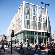 A busy urban street scene with a modern office building featuring large windows and a grid-like facade. Pedestrians cross at a marked crosswalk while cars wait at a traffic light. The clear blue sky and bright sunlight highlight the active city environment.