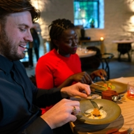 Two diners seated at a wooden table in a softly lit restaurant, eating a carefully plated dish with drinks beside them.