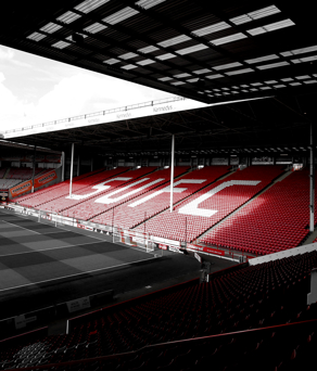 Interior view of a football stadium with red seating arranged around the pitch. Large white letters spelling ‘SUFC’ are visible on the main stand. The pitch is empty and partially shaded by the roof.