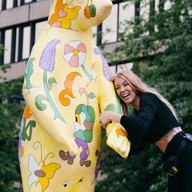 Large yellow bear sculpture decorated with colorful floral and abstract patterns, positioned outdoors near a modern building. A person is interacting with the sculpture, holding its arm