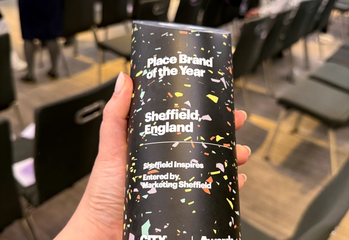 A hand holding a tall, cylindrical award with a colourful confetti‑style design. The text on the award reads “Place Brand of the Year – Sheffield, England – Sheffield Inspires – Entered by Marketing Sheffield – City Nation Place Awards 2025.” Rows of chairs and people mingling in the event space appear softly in the background.