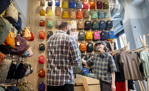Inside a retail store with a wooden display wall covered in colorful backpacks arranged in neat rows. Two people stand near a light wooden counter, one appearing to assist the other. To the right, racks display neatly hung t-shirts in neutral tones. The store has bright lighting and a modern, organized layout.