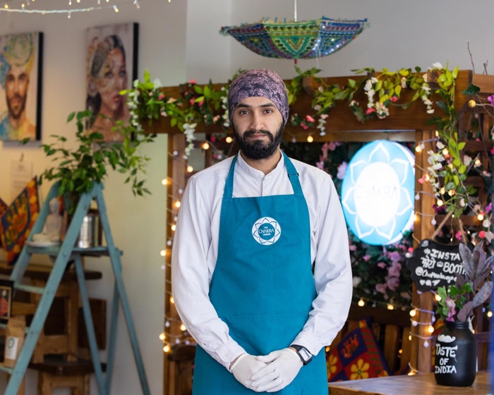 A person wearing a teal apron and white gloves stands inside a decorated shop or café. The background features colorful patterned cushions, hanging string lights, green plants, and a wooden display with flowers. A round illuminated sign and a chalkboard reading “Taste of India” are visible behind the person.
