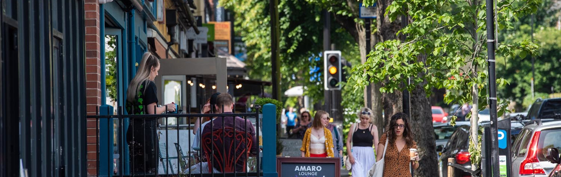 A row of shops and restaurants on a tree lined street. There are people walking up and down the pavement.