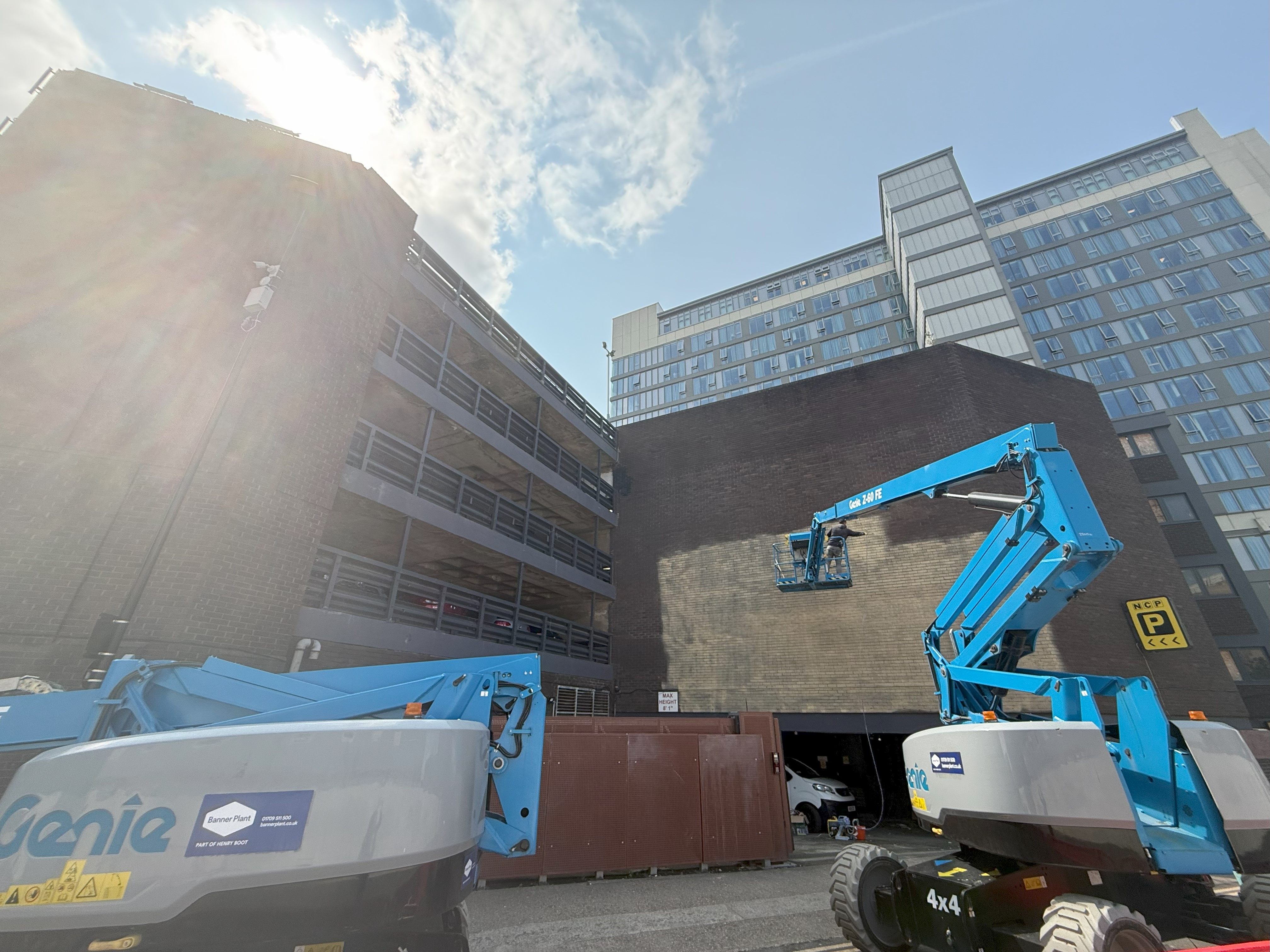An artist paints the base coat on a large angular brick wall from a blue cherry picker elevated in front