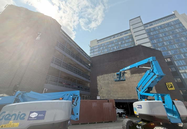 Construction scene in an urban area with two blue Genie boom lifts parked on a paved surface. One lift is extended toward a large blank brick wall, possibly for maintenance or mural work. Surrounding buildings include a multi-level parking structure on the left and a tall modern glass-fronted building on the right. A bright blue sky with scattered clouds and sunlight creates lens flare in the upper left corner.