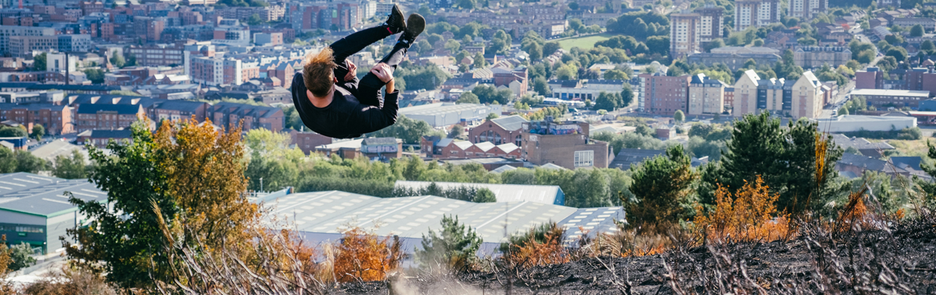 Person performing a mid-air flip on a hillside with burnt vegetation in the foreground, overlooking a cityscape with numerous buildings, trees, and distant hills under a partly cloudy sky.