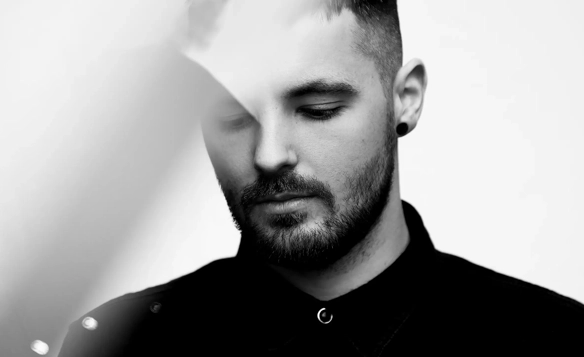 Close-up of a person wearing a dark collared shirt with metal buttons and a small hoop earring, standing against a plain light background.