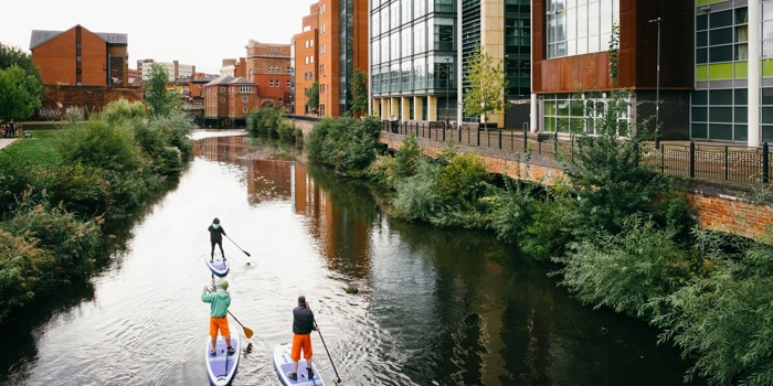 Three people are stand up paddle boarding on a canal that runs through a city centre.