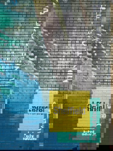 Close-up of a painted brick wall featuring part of a mural with blue and green water-like patterns. The word ‘PEACHZZ’ is written vertically in white letters on the right side. At the bottom, a yellow and green block of text reads ‘Sheffield Festival of the Outdoors .’