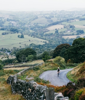 A scenic countryside view with rolling green hills and patchwork fields stretching into the distance. A narrow winding road runs through the foreground, bordered by a stone wall and patches of heather. A cyclist is riding along the road, surrounded by trees and shrubs. The background reveals a valley dotted with farms, hedgerows, and clusters of trees under an overcast sky, creating a tranquil rural atmosphere.
