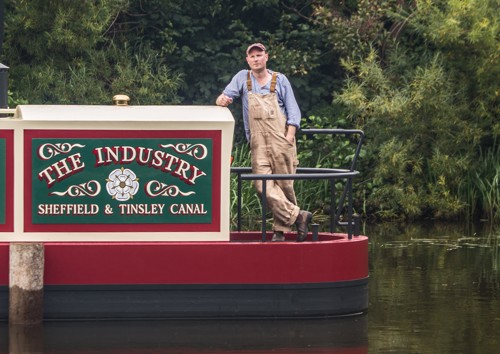 Person wearing light-colored overalls and a blue shirt leaning against a canal boat with a decorative sign reading ‘THE INDUSTRY Sheffield & Tinsley Canal,’ surrounded by green foliage.