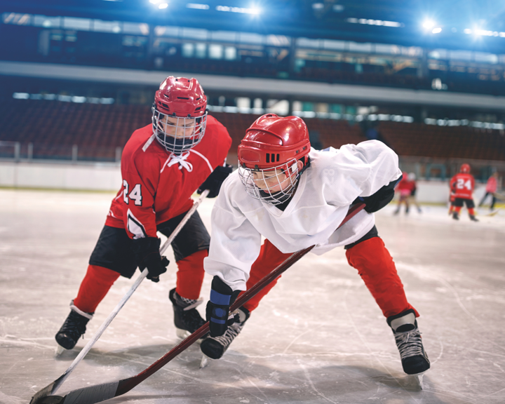 Two ice hockey players in full gear competing for the puck on an indoor rink. One player wears a white jersey and red pants, leaning forward with a stick on the ice, while the other in a red jersey closely defends. Additional players and seating are visible in the background under bright arena lights.