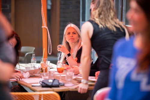 A group of people are gathered around a table in an outdoor dining area. The table holds drinks, glasses, and a small purse. One person with long light-colored hair is seated, holding a drink and facing another person who is standing with their back to the camera. The setting includes chairs, tables, and part of a building structure, with clear pathways and seating arrangements that support physical accessibility.
