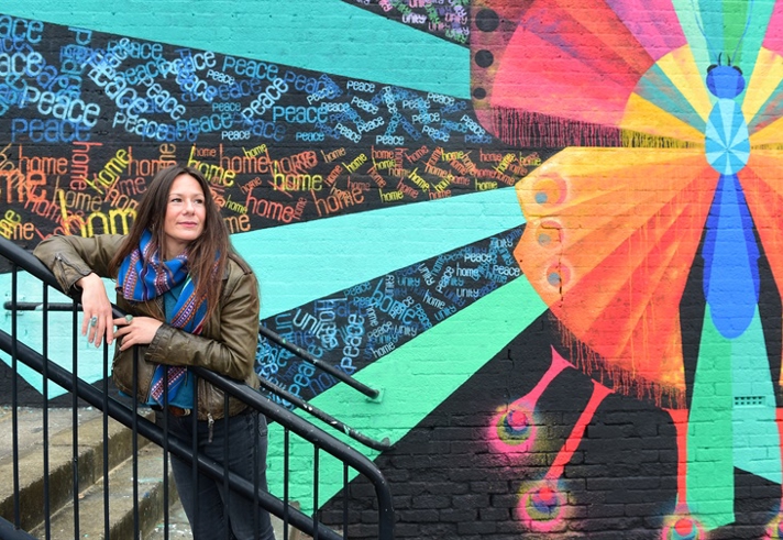 A woman leans on the handrail of some steps, in front of a mural 