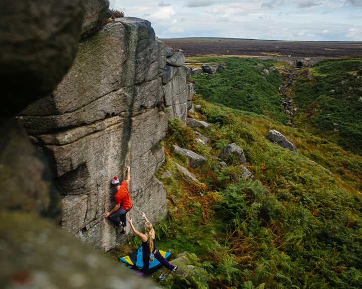 Two people are climbing up a rock face. They are using climbing gear and have a safety mat on the ground.