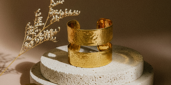 Elegant wide gold cuff bracelet with hammered texture, showcased on stacked stone display with geometric gold accents and dried foliage in soft lighting