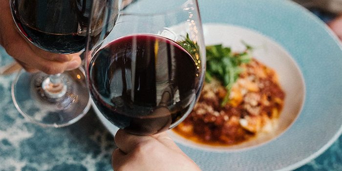 Two wine glasses clinking above plates of food.