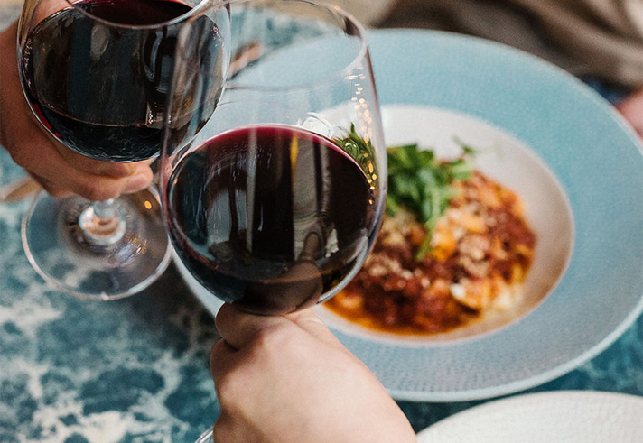 Two wine glasses clinking above plates of food.
