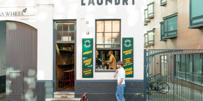 Exterior of a white-painted building with the word “LAUNDRY” in bold black letters above two windows and a doorway. The entrance is open, revealing a rustic interior with bar stools and a counter. Green signs with yellow text advertise coffee and good vibes. A person stands outside on the pavement near a sandwich board, while another is visible inside by the counter. The setting is urban, with a gated area and bicycles to the right.