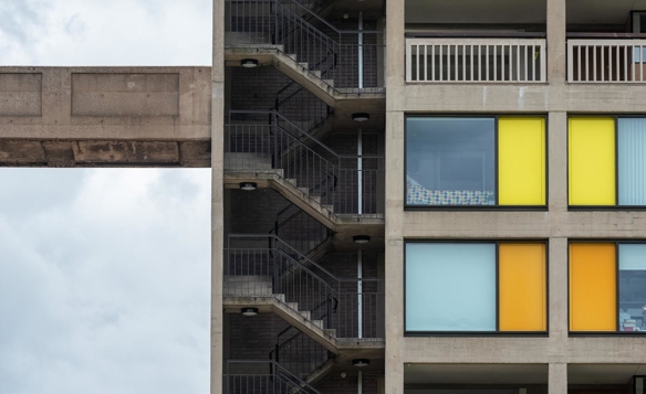 Close-up view of a modern concrete apartment building with an external zigzag staircase on the left and colorful window panels on the right in shades of yellow, orange, and blue. The structure includes connecting walkways and balconies, set against a cloudy sky.
