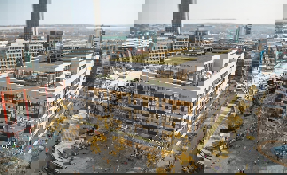 Aerial view of a modern multi-storey building labeled ‘COLE STORE’ with large glass windows and a green rooftop terrace. The building is surrounded by wide pedestrian areas with trees and people walking. In the background, there are high-rise office buildings, construction scaffolding on the left, and a mix of older and newer architecture under a hazy sky.