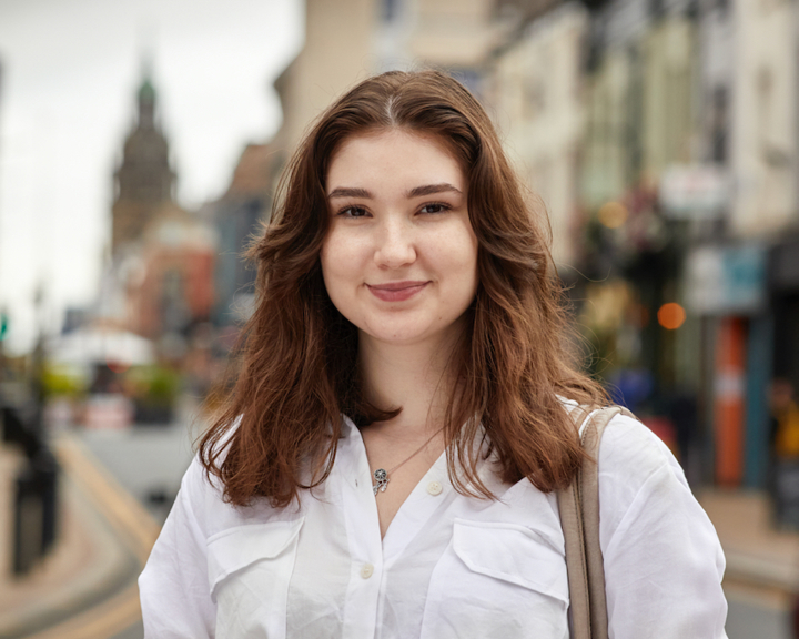 A person wearing a white button-up shirt and black belted trousers is standing on a city street with buildings lining both sides. The individual has a beige shoulder bag and is positioned in the foreground, while the background shows a slightly overcast sky and a distant clock tower. The street features double yellow lines and appears to be in an urban area with shops and pedestrians.