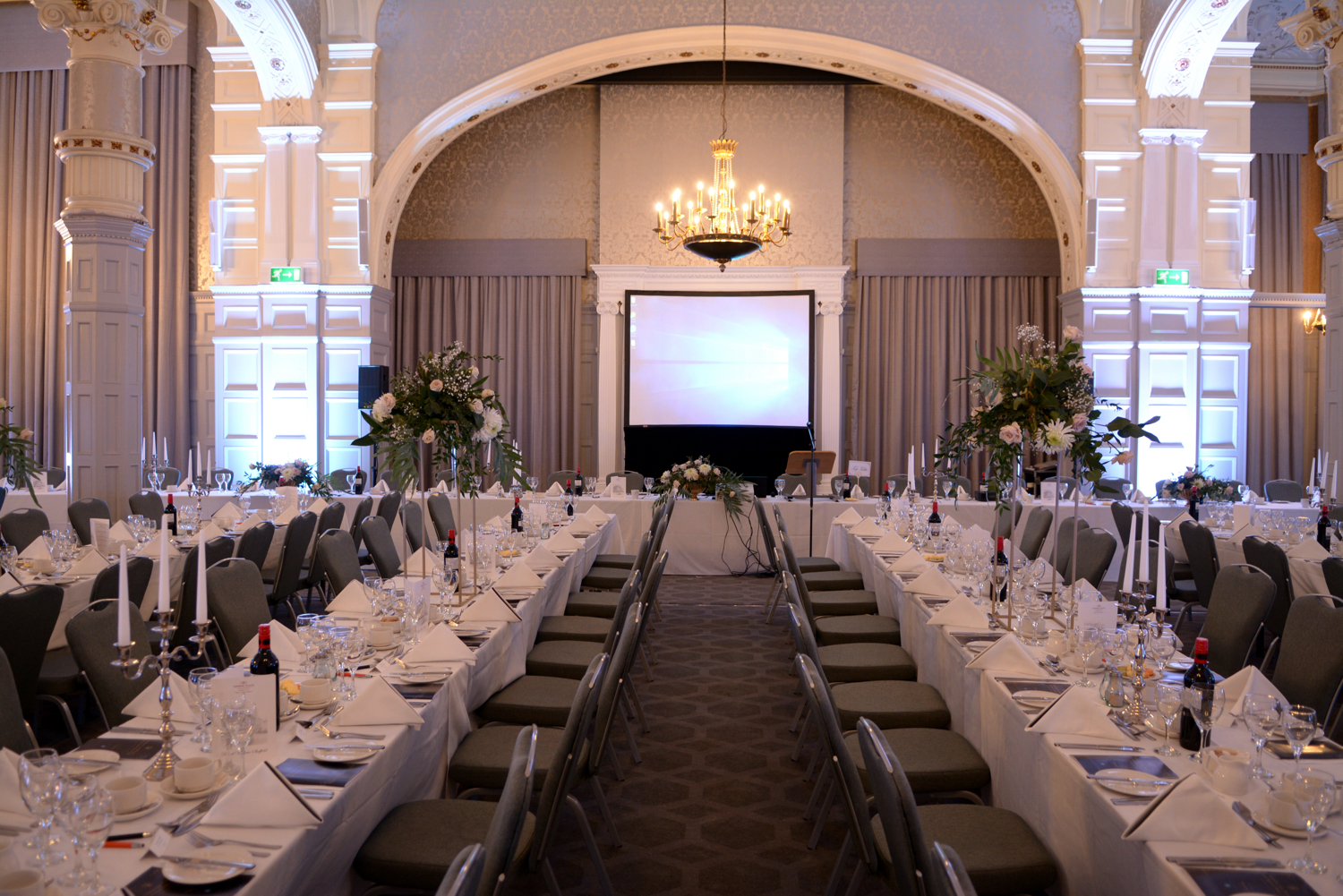 Crowne Plaza Royal Victoria - Elegant banquet hall with high arched ceilings, ornate detailing, and a chandelier. Long tables are set with white linens, glassware, candles, and floral centerpieces, facing a projection screen at the front.