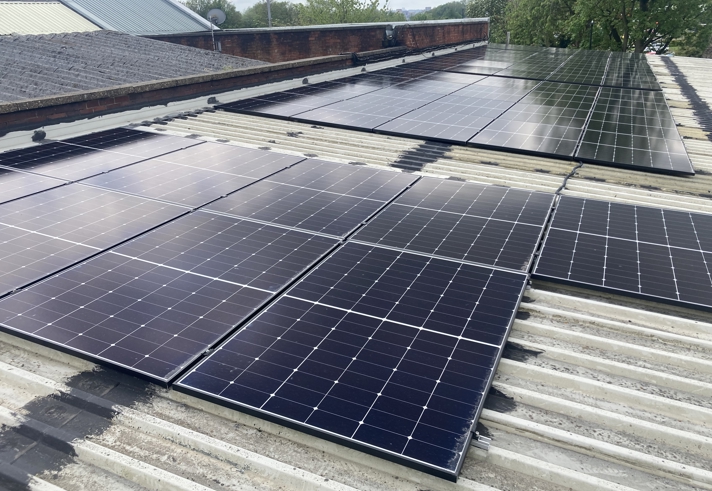 Rows of black solar panels installed on a corrugated metal rooftop, with additional panels visible further back. Surrounding elements include a brick wall, part of a sloped roof, and green trees in the background under a partly cloudy sky.