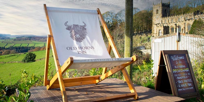 A large wooden deckchair printed with the Old Horns Inn logo stands on a raised platform overlooking rolling green countryside, with a historic stone church visible in the background under a cloudy sky.