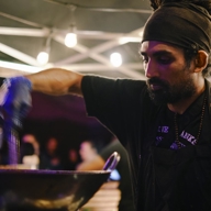 A person wearing a dark apron and head wrap is cooking at an outdoor food stall under a canopy with string lights. The individual is stirring food in a large wok, and bottles of condiments are visible in the background along with blurred figures and warm lighting.
