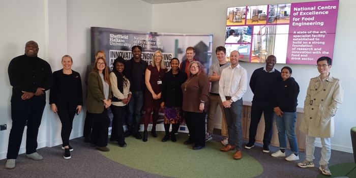 A group of twelve people standing in a semi-circle inside a modern meeting room with grey carpet and green accents. Behind them is a large banner displaying “Sheffield Hallam University” and “National Centre of Excellence for Food Engineering.” On the right wall, there is a digital screen showing images and text about the centre. The room has white walls, a ceiling light, and a few chairs visible on the side.