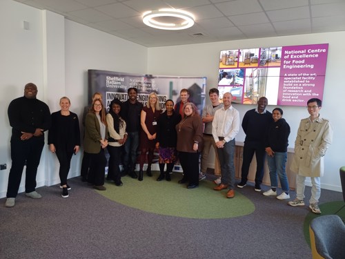 A group of twelve people standing in a semi-circle inside a modern meeting space with grey carpet and green accents. Behind them is a large banner displaying “Sheffield Hallam University” and “National Centre of Excellence for Food Engineering.” On the wall to the right, there is a digital screen showing images and text about the centre. The room has white walls, a ceiling light, and contemporary chairs arranged along the side.