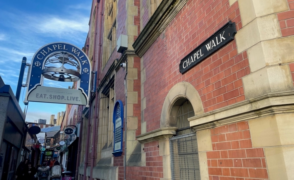 Narrow pedestrian walkway called Chapel Walk, lined with red brick buildings featuring stone detailing. A black sign reading ‘Chapel Walk’ is mounted on the wall, and a decorative hanging sign with the same name and the words ‘Eat. Shop. Live.’ is visible above the entrance. Several smaller shop signs and people walking can be seen further down the alley under a bright blue sky.